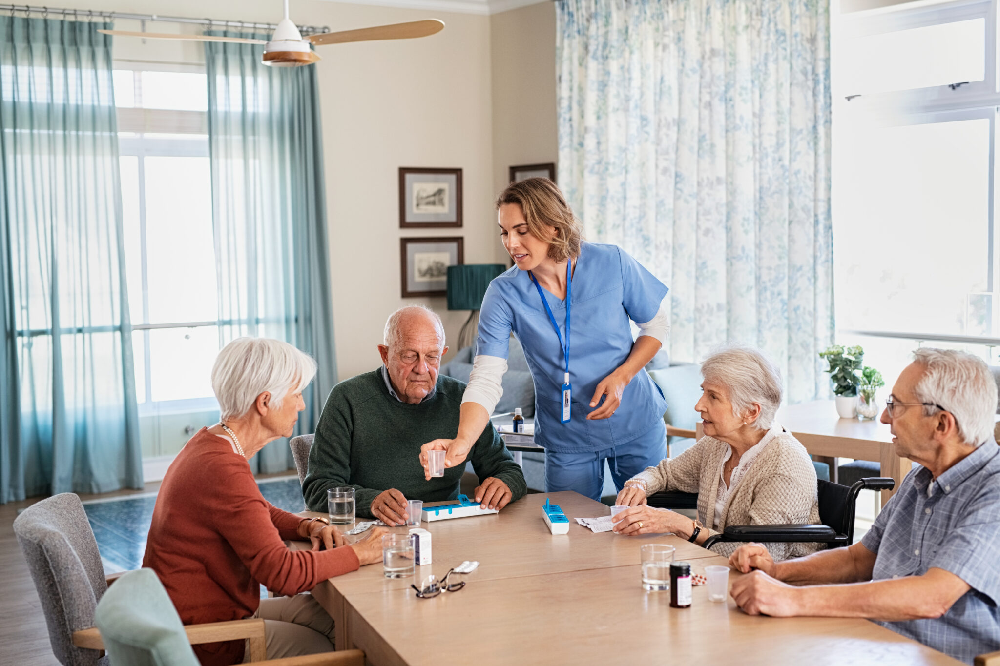 Young caregiver in uniform giving medicine to group of senior men and women in care centre. Female nurse giving daily pills to old woman after lunch in nursing home. Experienced doctor giving medicine to senior people at care facility. Assisted living costs