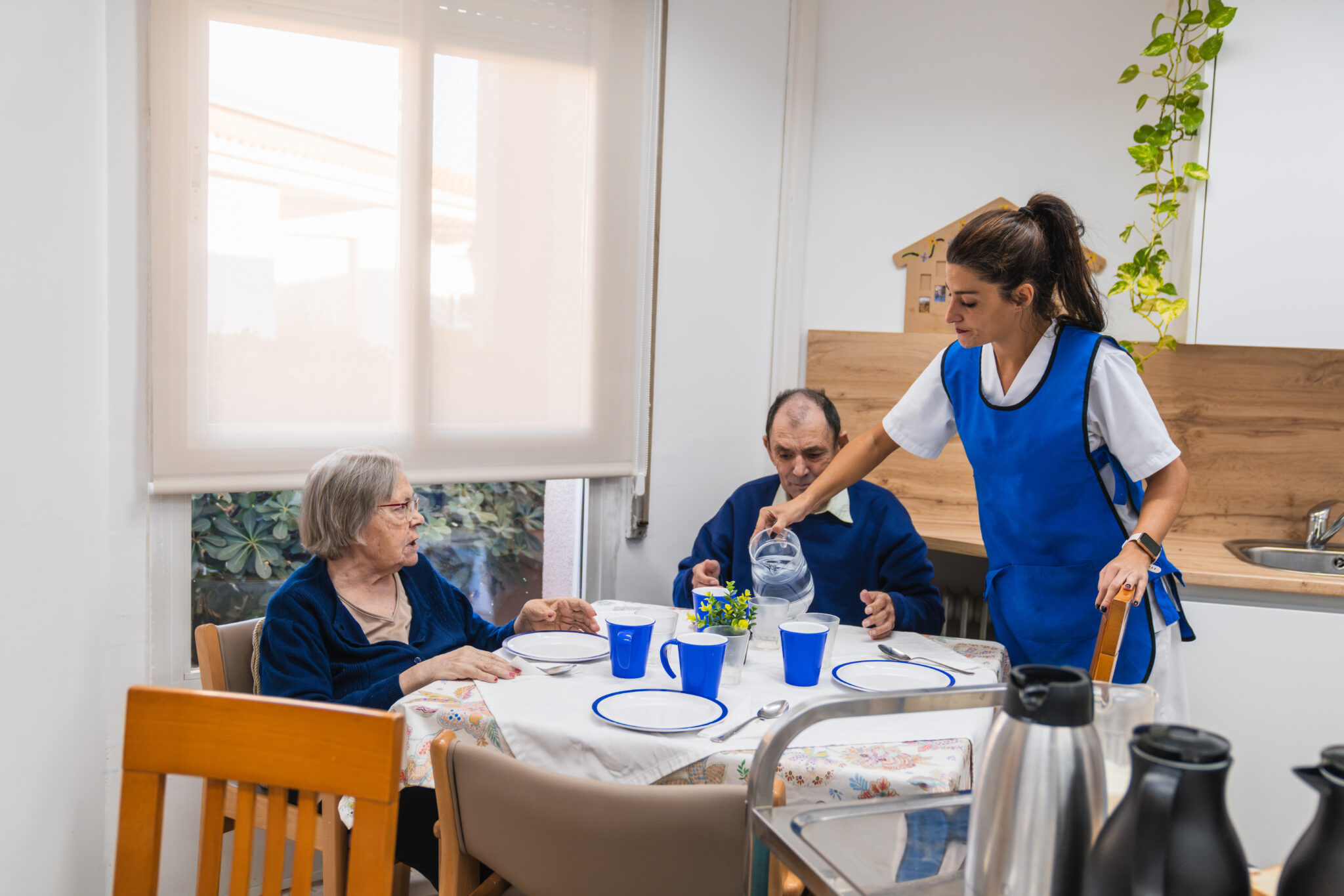 Caregiver in blue uniform serving water to elderly residents at a nursing home dining table, fostering a caring and supportive environment