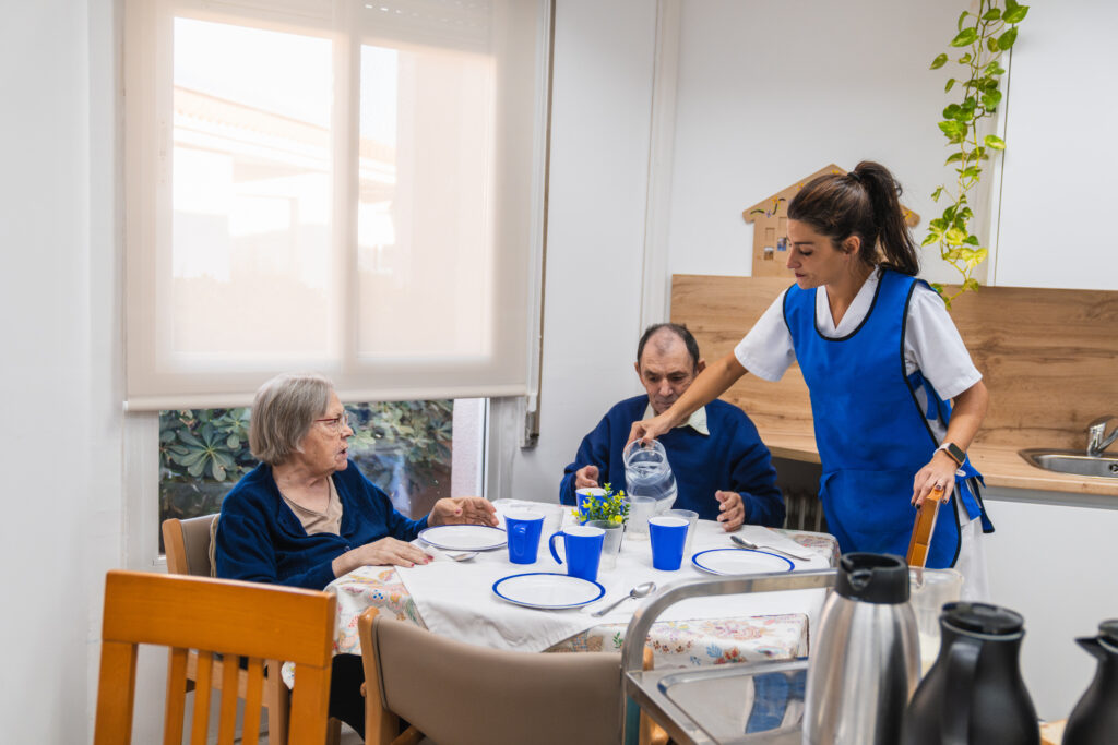 Assisted Living Levels of Care: Caregiver in blue uniform serving water to elderly residents at a nursing home dining table, fostering a caring and supportive environment