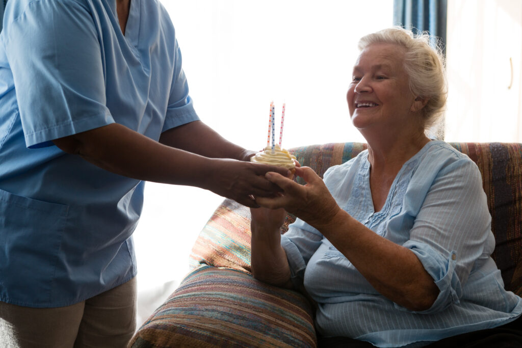 Assisted Living Questions to Ask: Happy senior woman looking at cup cake with candle held by nurse in nursing home