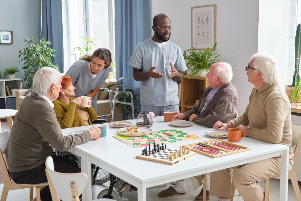 Activities in Assisted Living Facilities: Group of senior people playing board game at the table and talking to medical staff in the room of nursing home