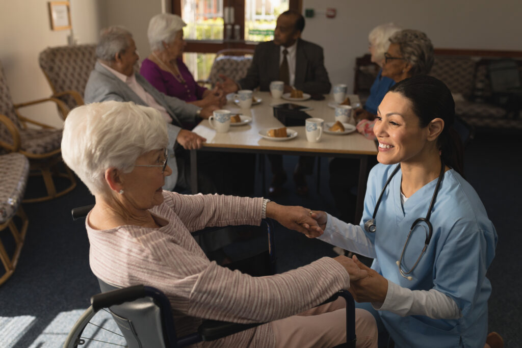 Side view of happy female doctor holding hands and talking with disable senior woman at nursing home