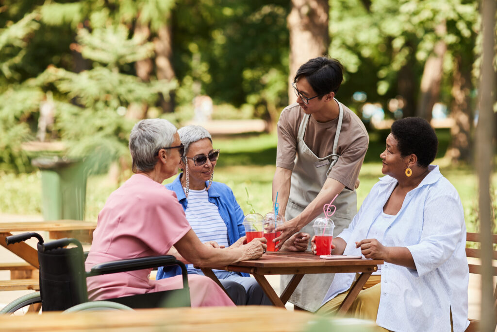 Portrait of young Asian waiter serving drinks to group of senior women at outdoor cafe assisted living communities
